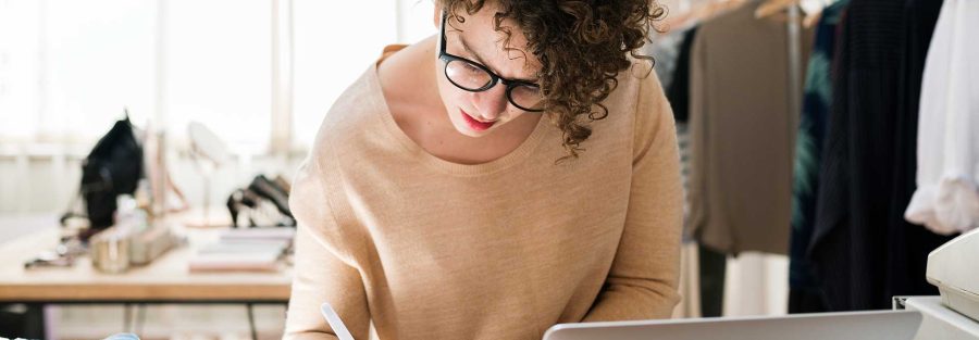 Woman writing in a notebook with a laptop in front of her